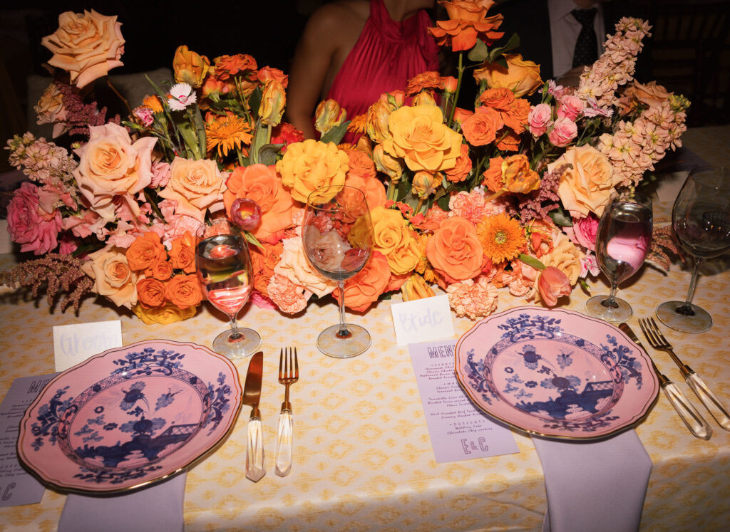 Spring wedding tablescape to match the garden wedding aesthetic with yellow and orange flower arrangements, pink and purple glassware, and a yellow patterned tablecloth.